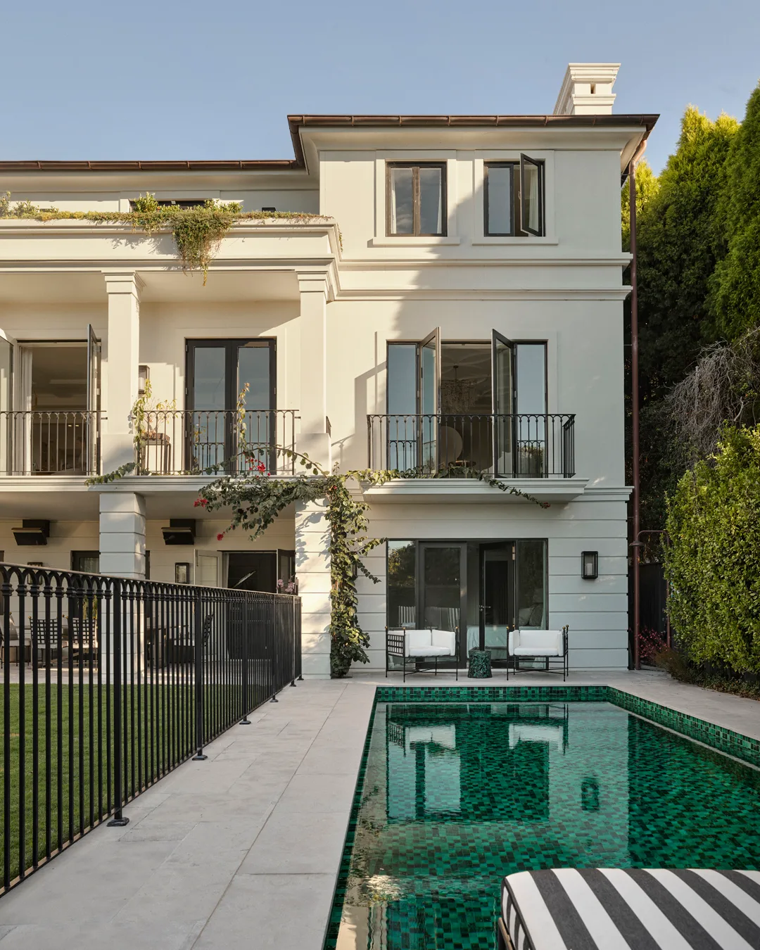 A three-story beige house with wooden window panelling, a green-tiled pool and black balustrade and grass