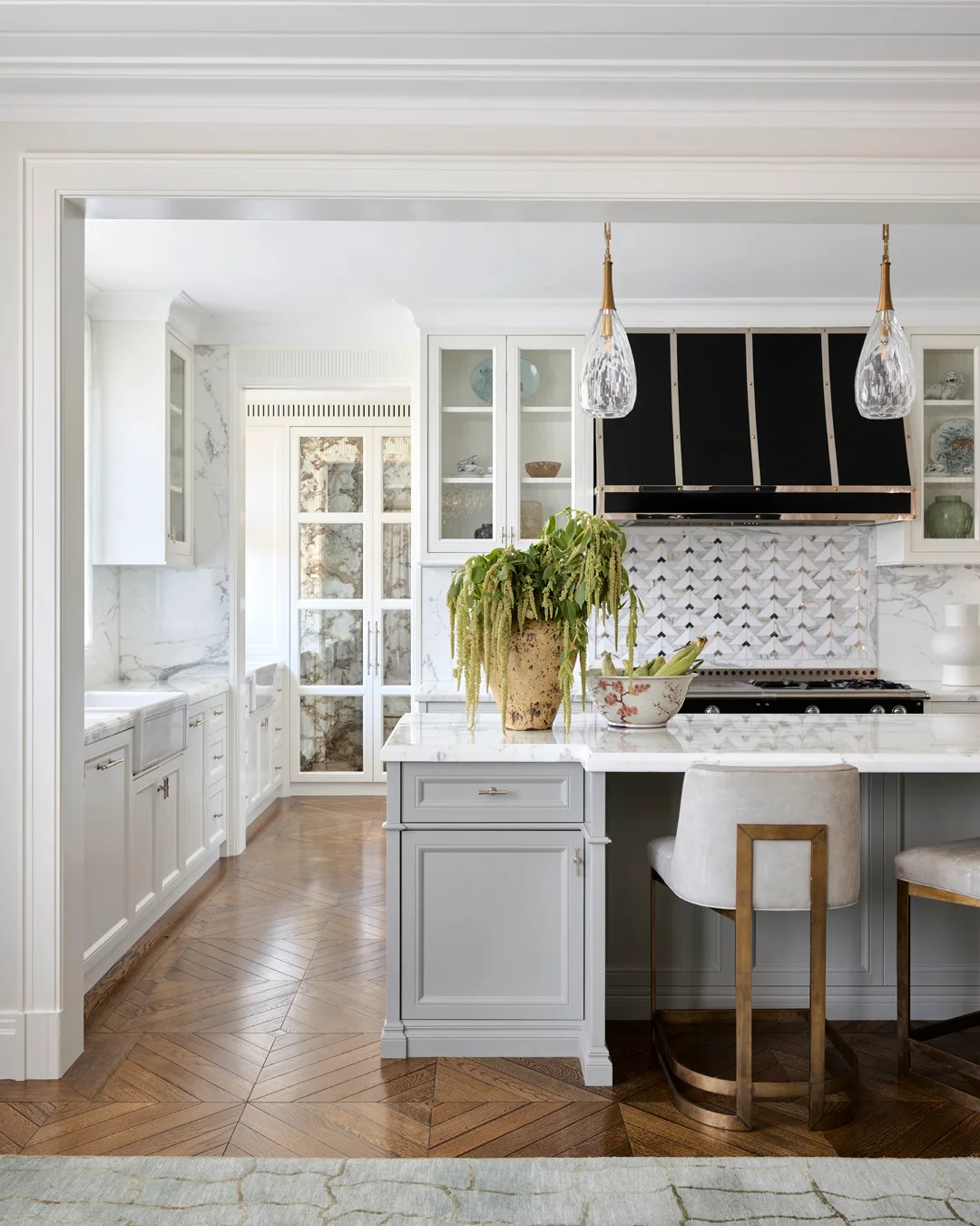 A marble and grey kitchen with black range hood, plush modern stools, timber floors, bowl of bananas and a vase with greenery