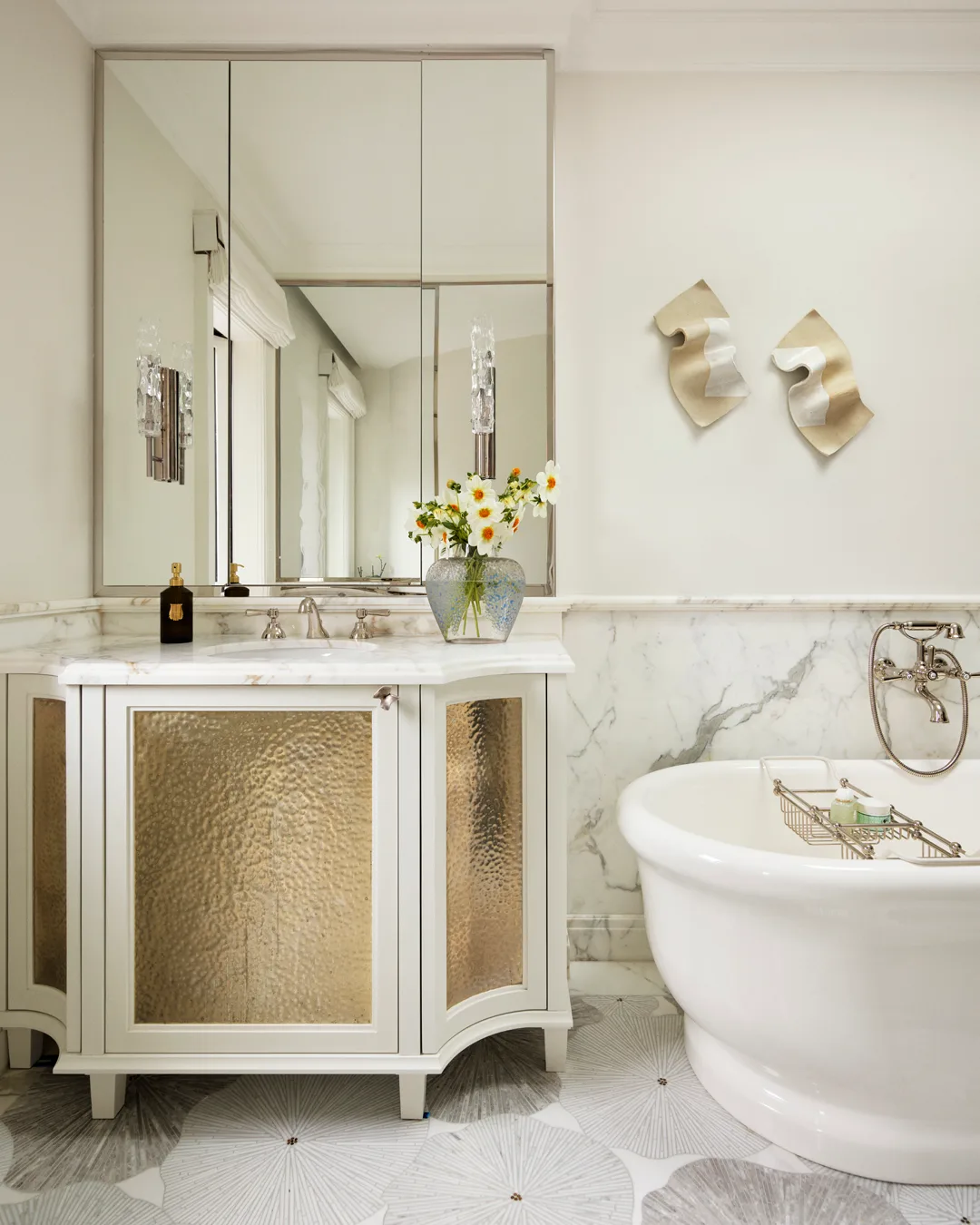 A bathroom with white and gold basin, white bathtub, glass vase of flowers, abstract artwork and large mirror