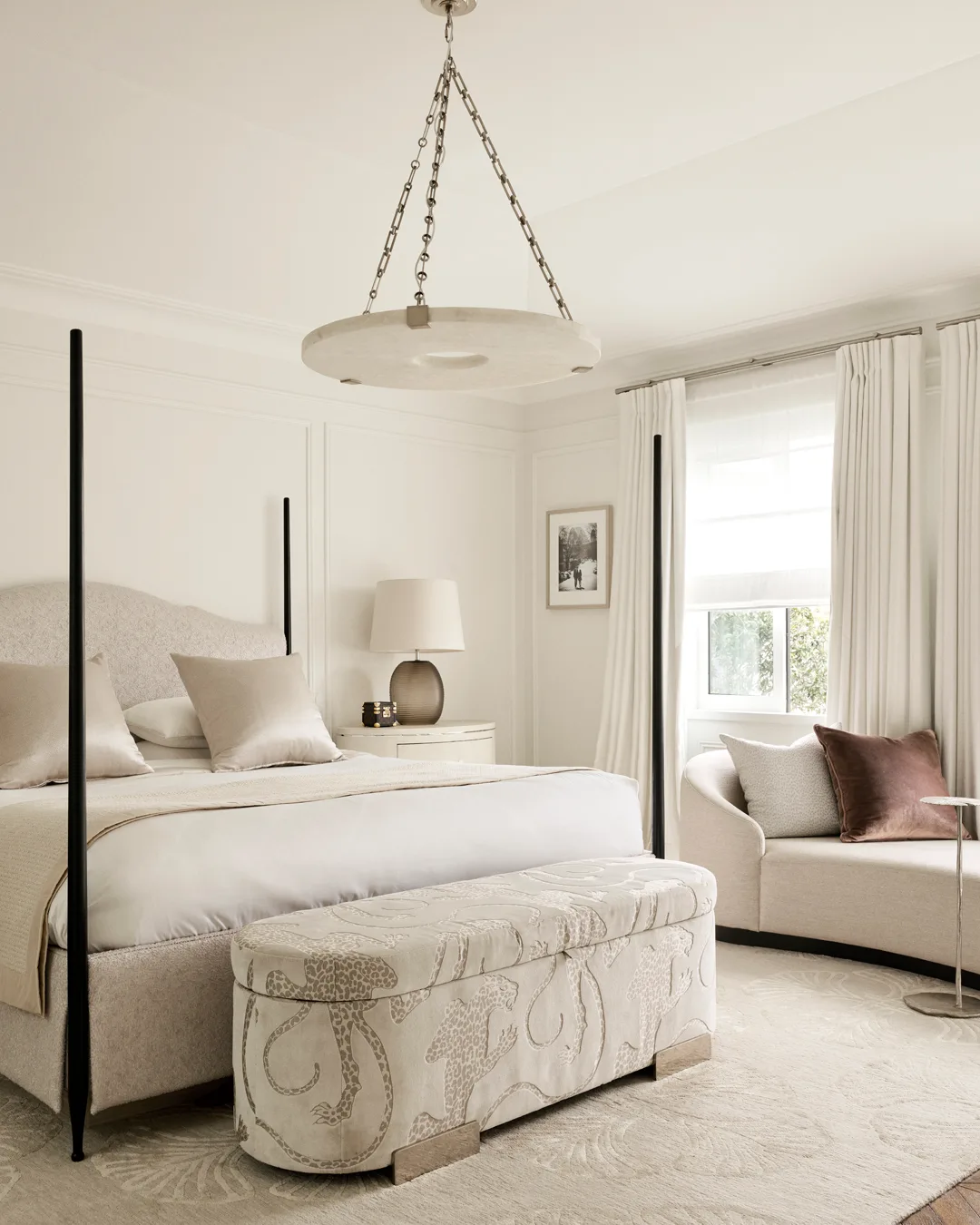 A bedroom with natural bedding, white, cheetah-print footstool, modern chandelier, cream sofa bed and cream certains