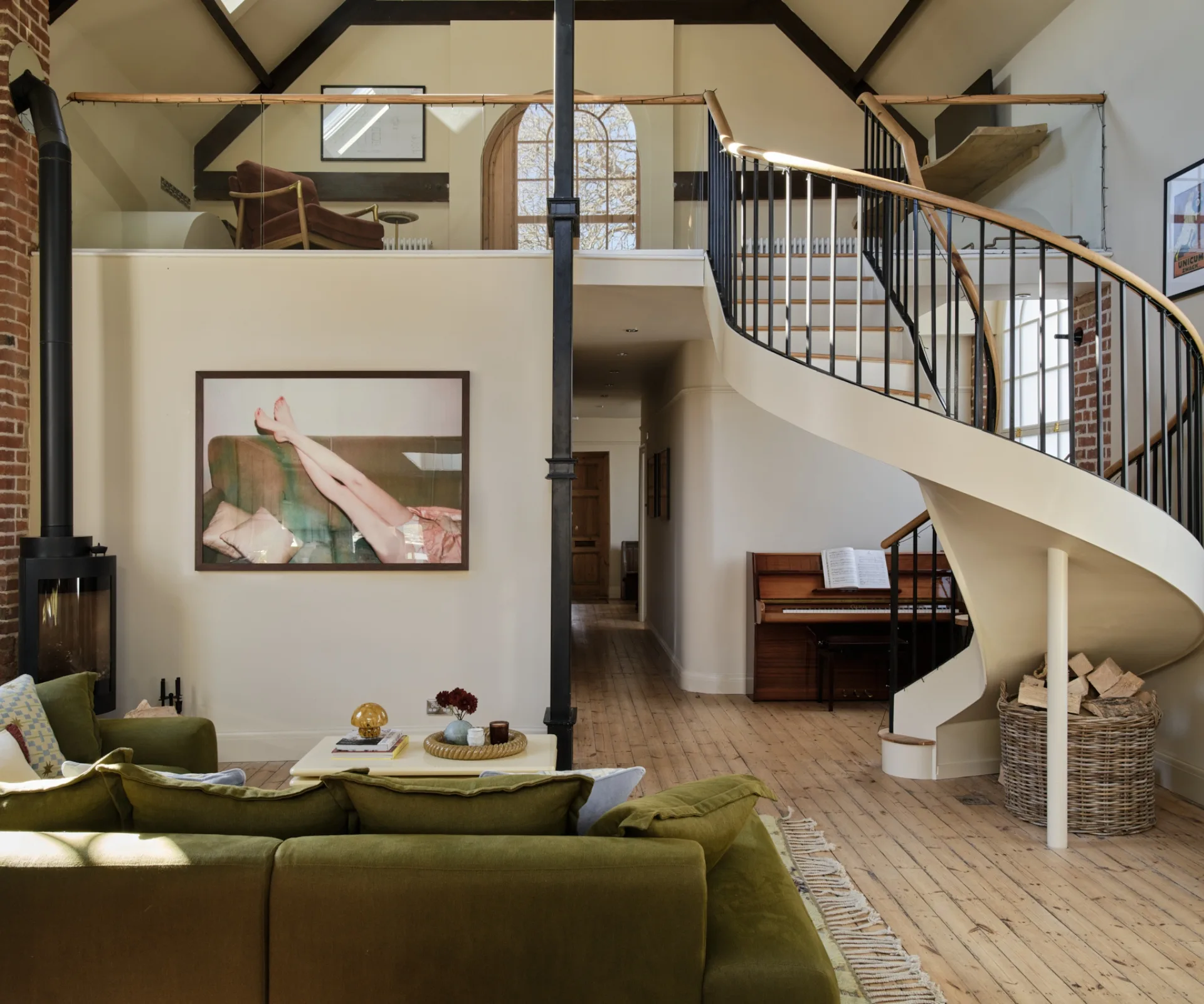 Double-height living space inside a converted chapel with an olive-green modular sofa, modern fireplace, curved white staircase, and exposed brick detailing.