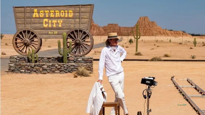A man wearing a white outfit standing in the desert with a director camera