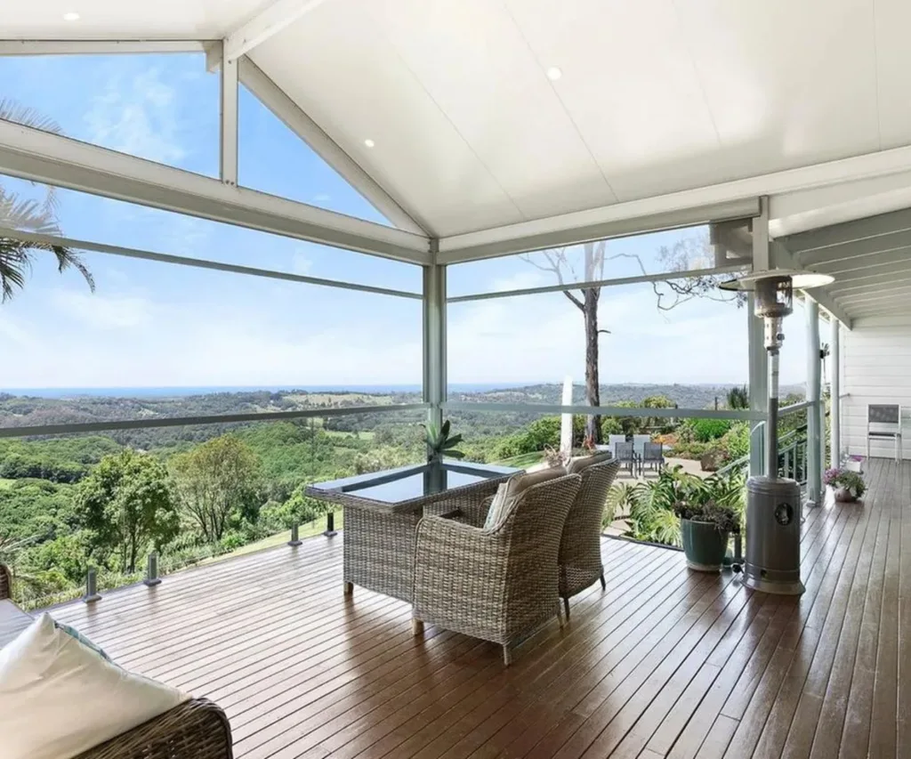 Covered deck with wicker furniture overlooking green hills and distant ocean view, under a clear blue sky.
