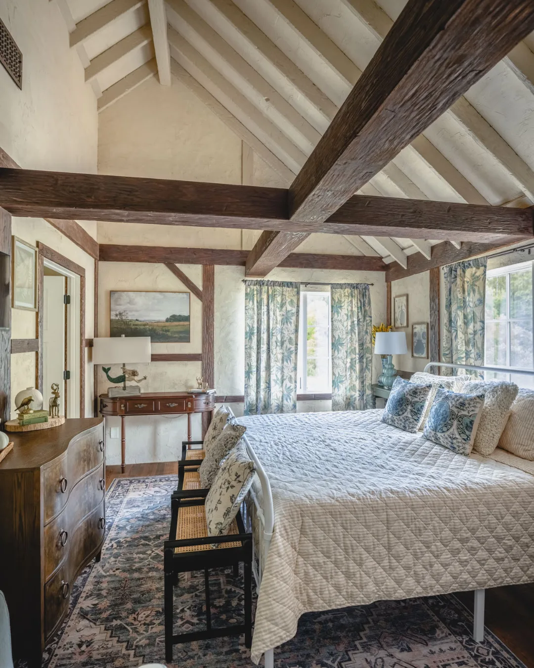 A cottage bedroom with a paisley bed cover, timber beams, Persian rug and high cream ceiling. 