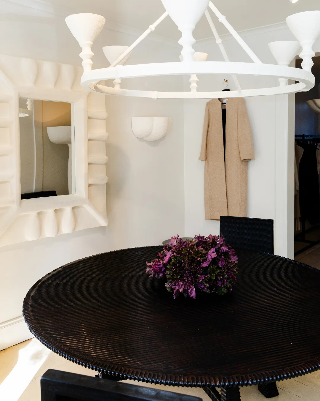 A timber dining table against white walls, with white framed mirror, beige coat hanging on the wall and ceramic chandelier