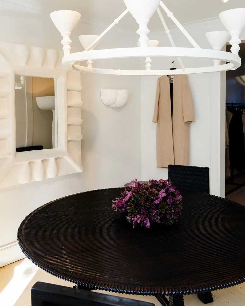 A timber dining table against white walls, with white framed mirror, beige coat hanging on the wall and ceramic chandelier