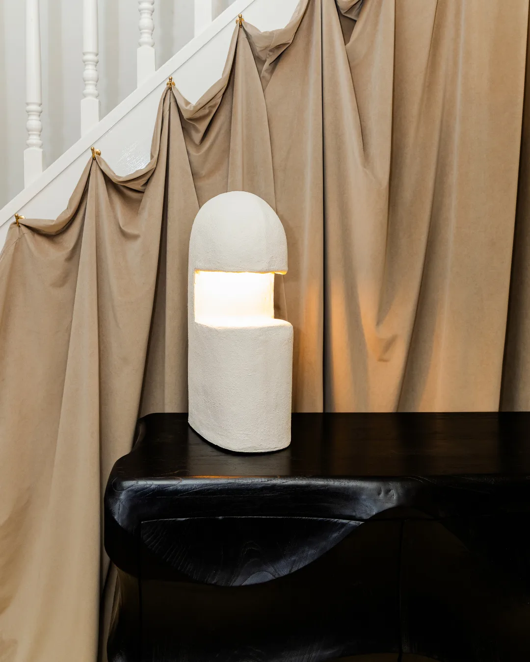 A modern white lamp sits on top of a black timber sideboard and in front of a beige curtain hung on a stairwell balustrade