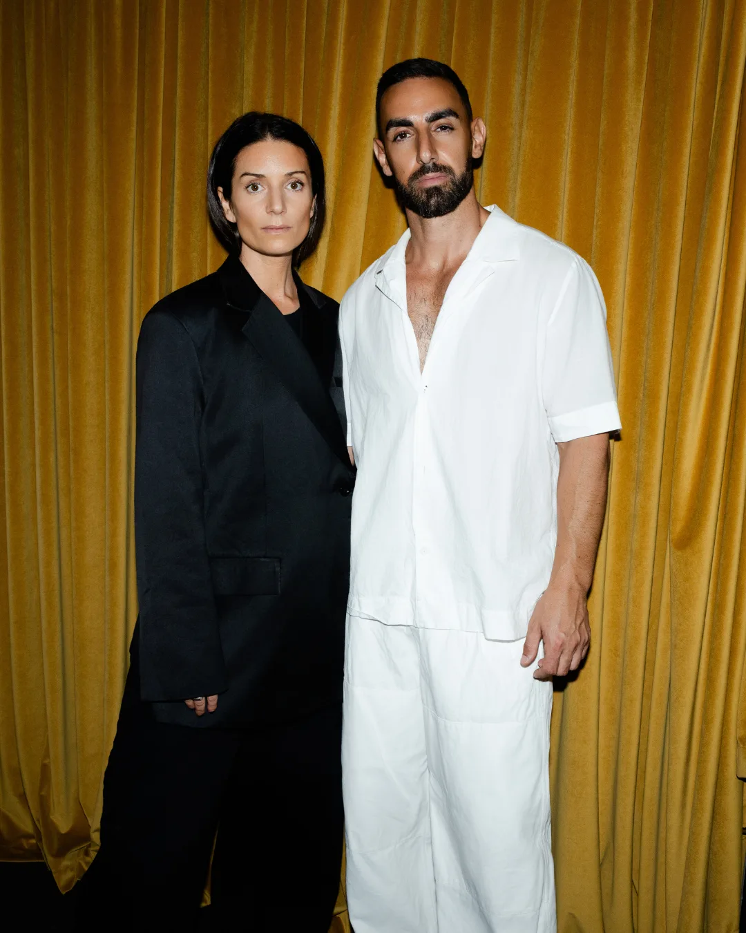 A man dressed in white and a woman dressed in black stand in front of a mustard, velvet curtain