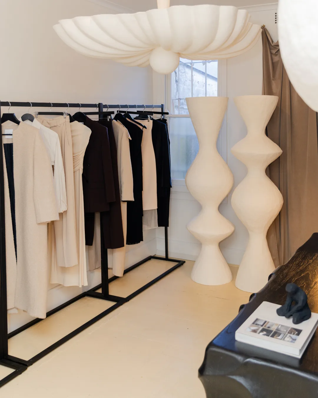 A rakc of monochromatic clothing hanging on a rack, with two ceramic floor lamps and a ceramic chandelier 