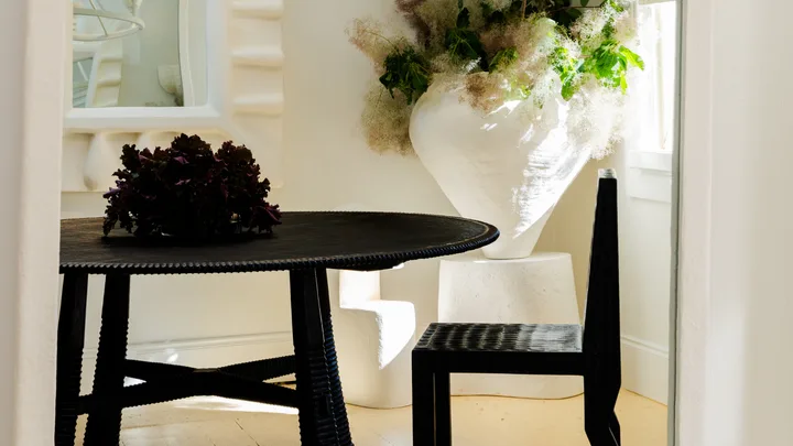 A dining room with white walls, giant white flower vase with flowers, a white ceramic chandelier, dark timber table and chairs