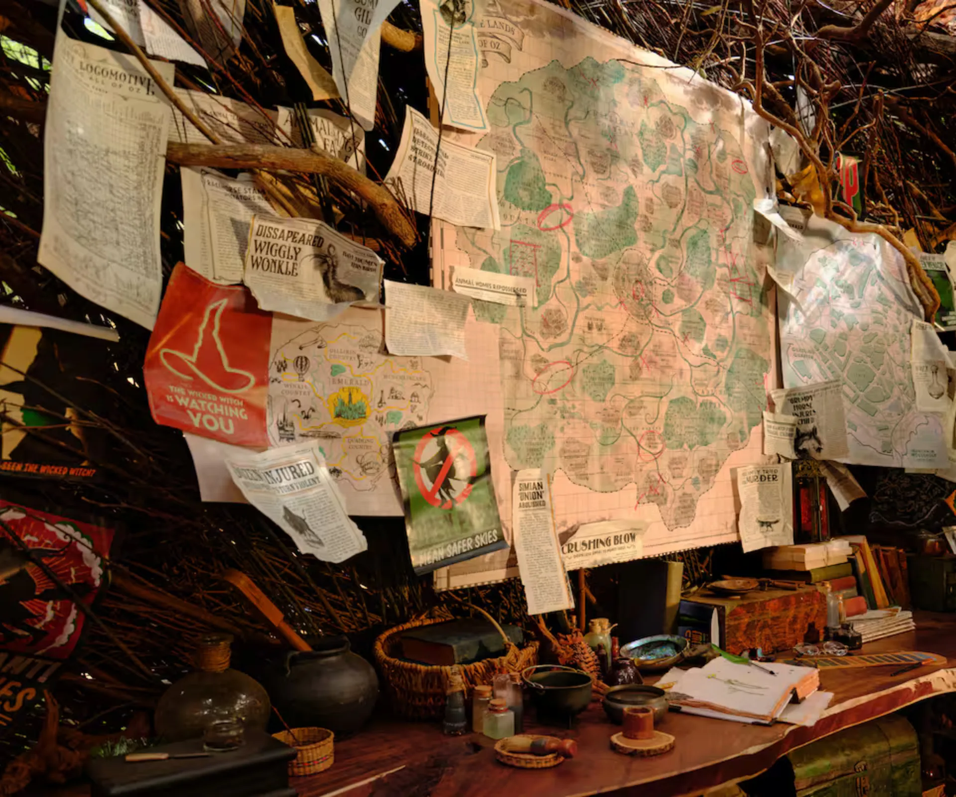 A map and signage pinned to a wall covered in vines above a desk, with paper, books and pots