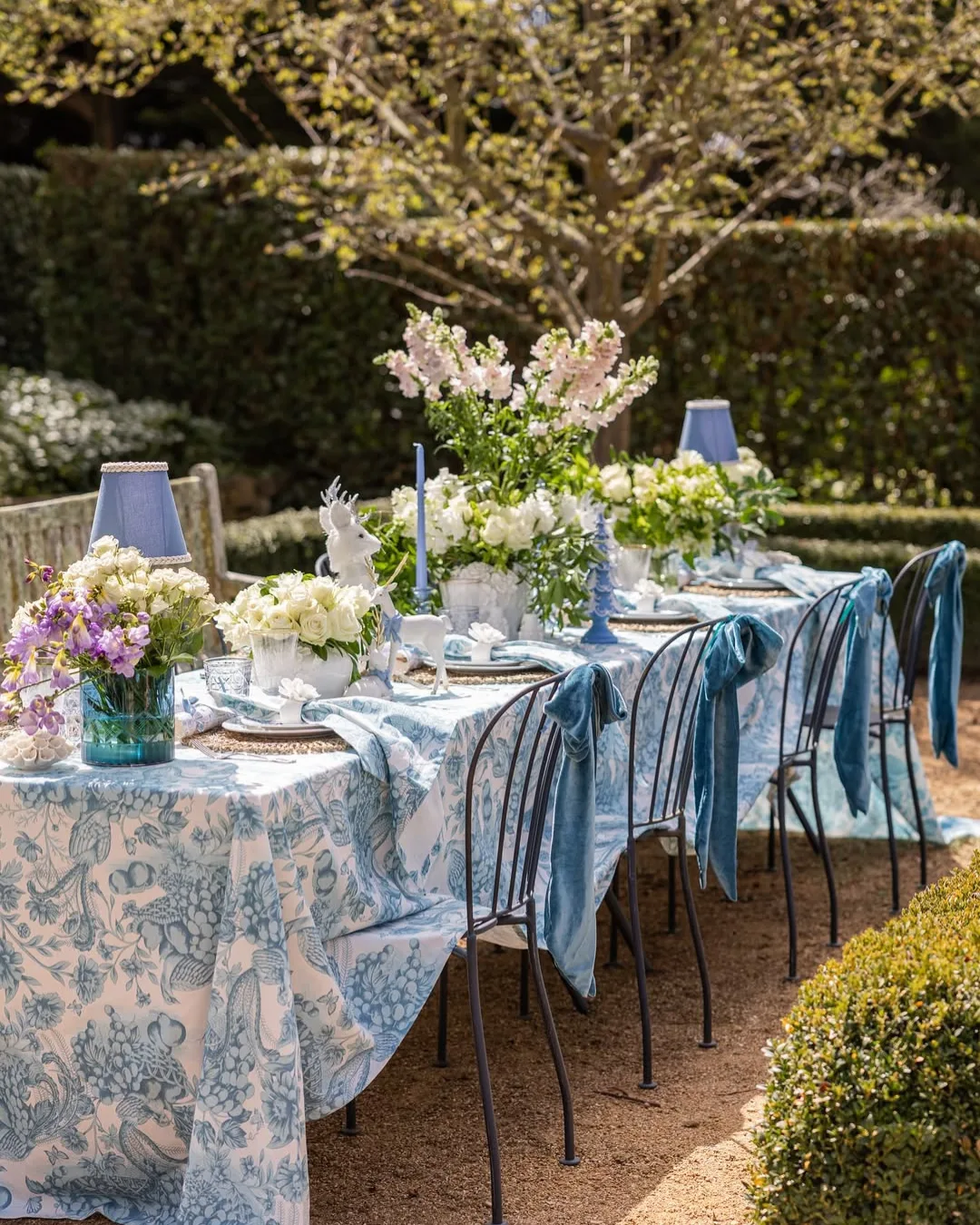 A table set with a blue and white pattern tablecloth, flowers, velvet blue bows and tableware in a garden