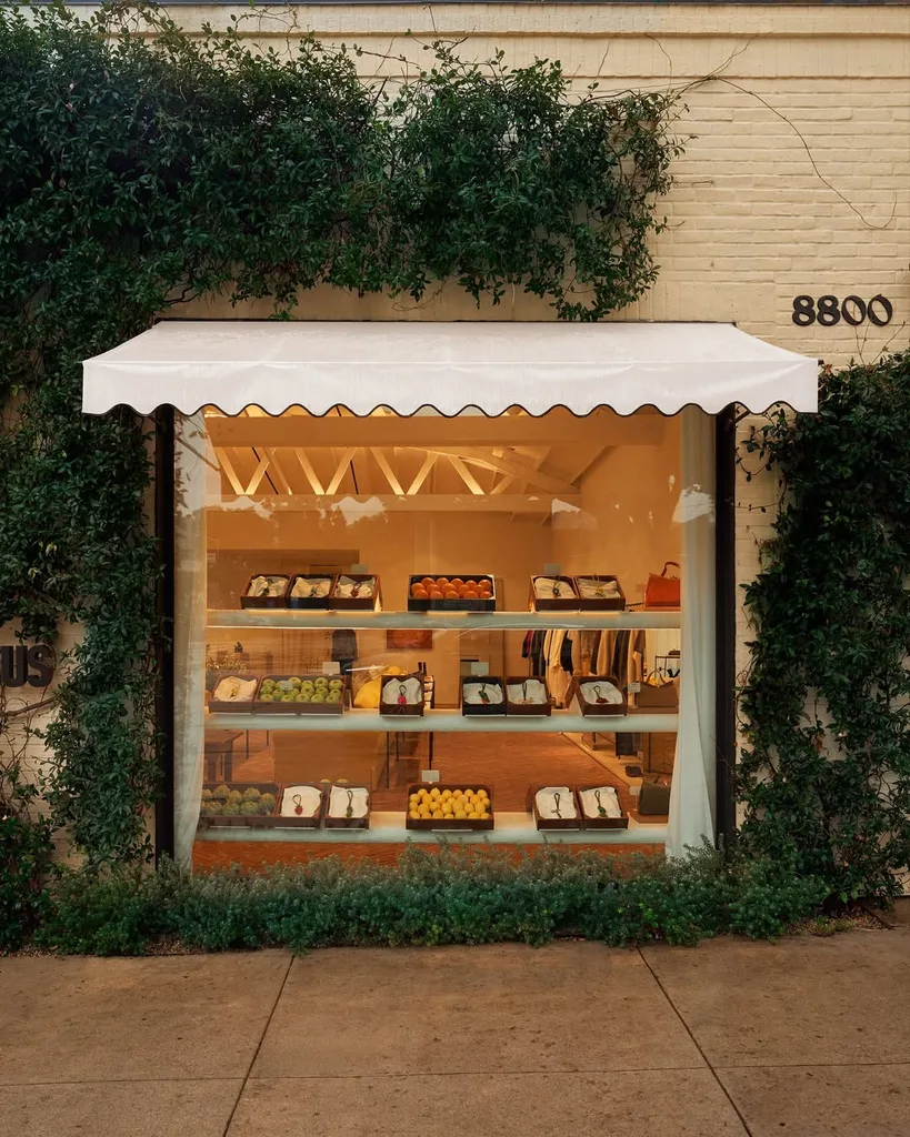 Le Valérie Jacquemus handbag launch with a shop front with white scalloped awning, glass window with crate of fruit and clothes and greenery