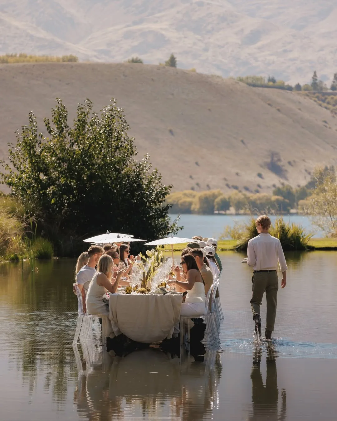 An outdoor setting with a table in a body of water, mountains, people eating with umbrellas
