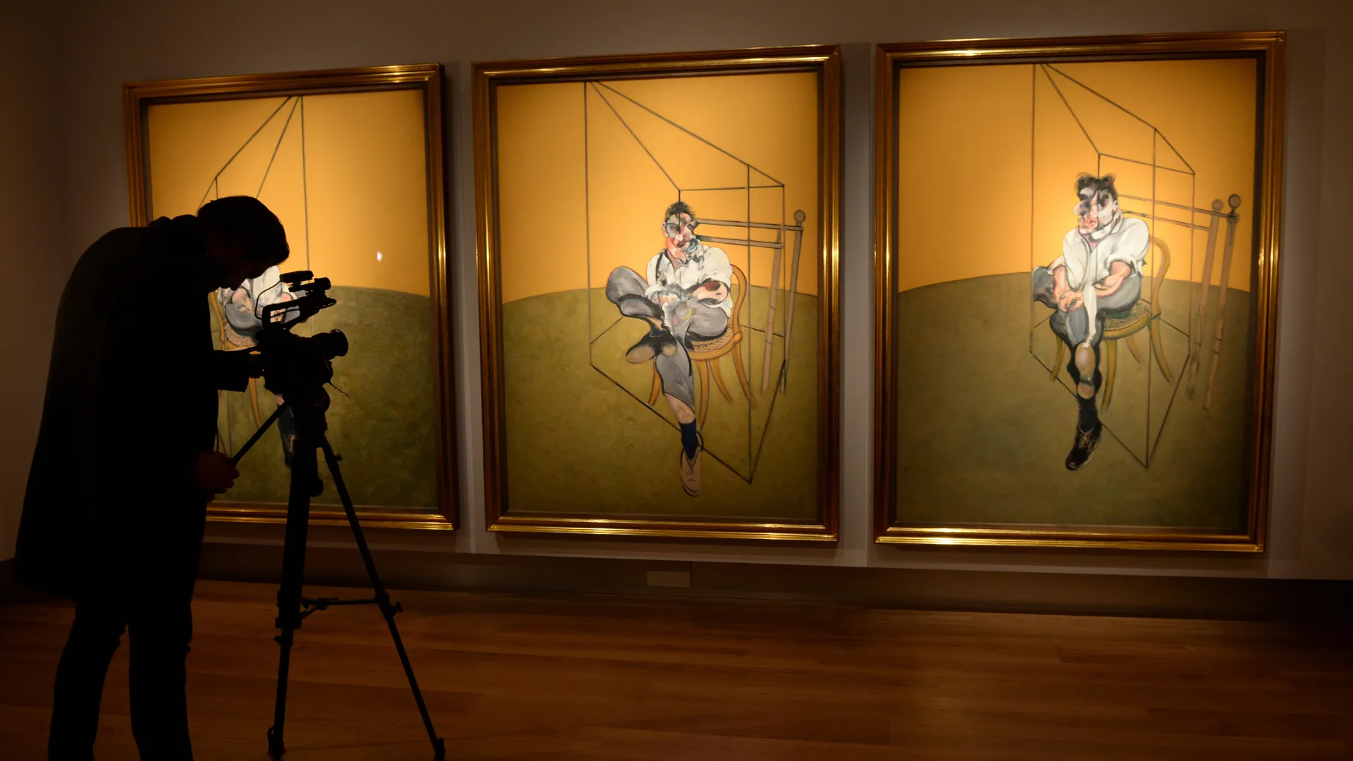 A man taking a photo of three painting in gold frames ide by side