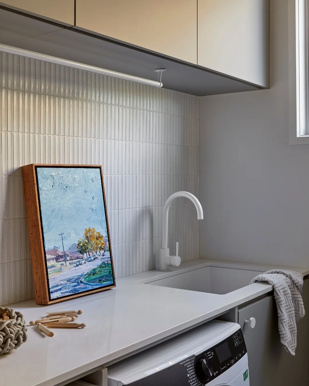 A landscape painting sits on the laundry bench against a tiled backsplash. 