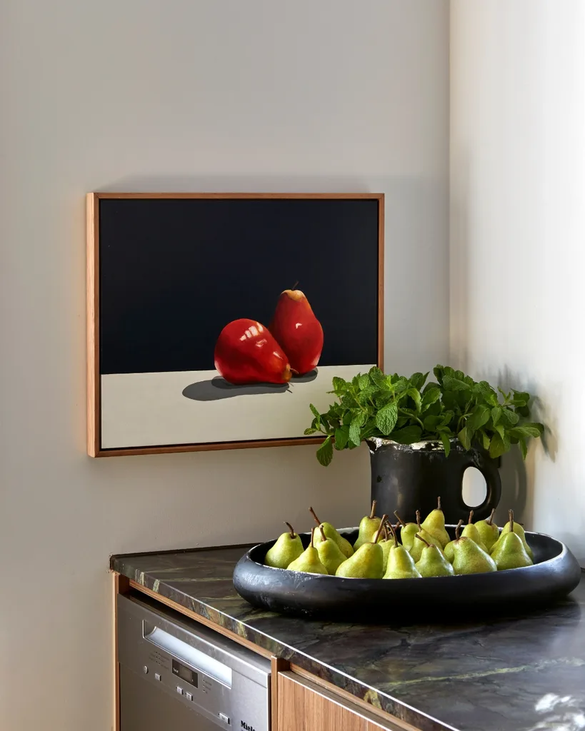 Still life artwork by Nell Symonds depicting a pair of red pears against a black background hangs in a kitchen above a bowl of green pears.