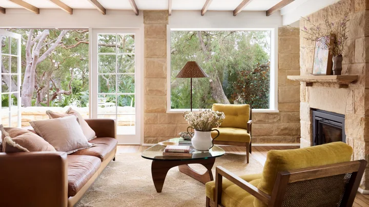 Coastal living room with sandstone walls, tan sofa, mustard chairs, and large windows overlooking lush greenery.