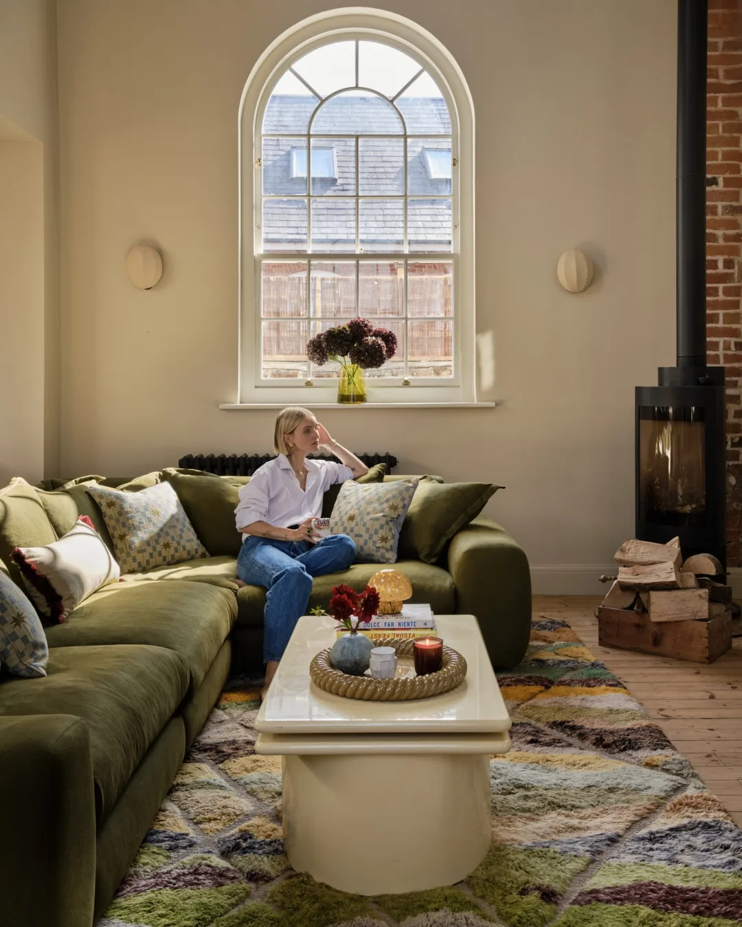 Polly Sayer sits on a green L-shaped sofa under original arched windows in living room. 