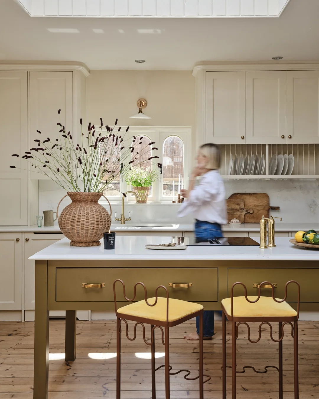 Neutral country kitchen with olive island, brass details, wicker vase and sculptural bar stools under a skylight in a renovated English chapel.