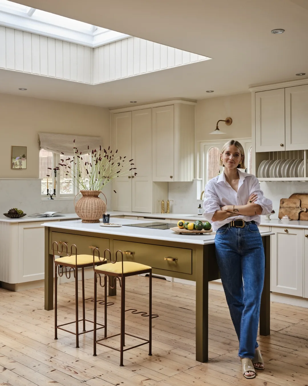 Polly Sayer stands in her neutral country kitchen with olive island, brass details, wicker vase and sculptural bar stools under a skylight in her renovated English chapel.