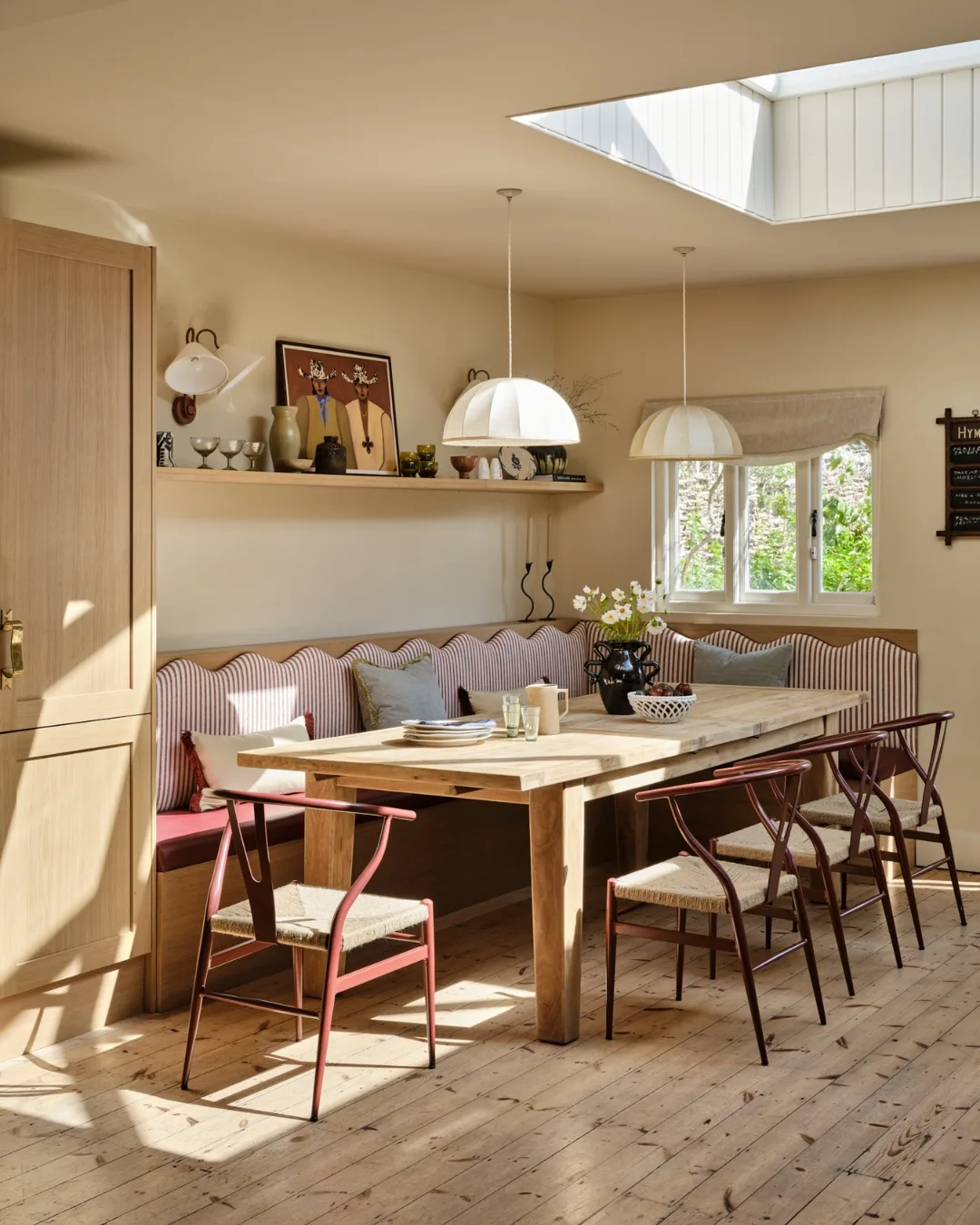 Warm, light-filled dining nook in a renovated English chapel featuring a striped red-and-white scalloped banquette, oak table, and red wishbone-style chairs under two paper pendant lamps. A skylight above bathes the space in natural light. 