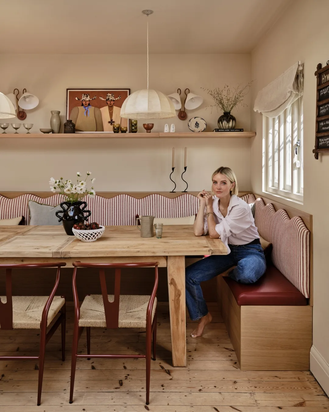 Fashion director Polly Sayer sits in her dining nook with striped banquette, oak table and wall shelf of ceramics - timeless English country kitchen inspiration.