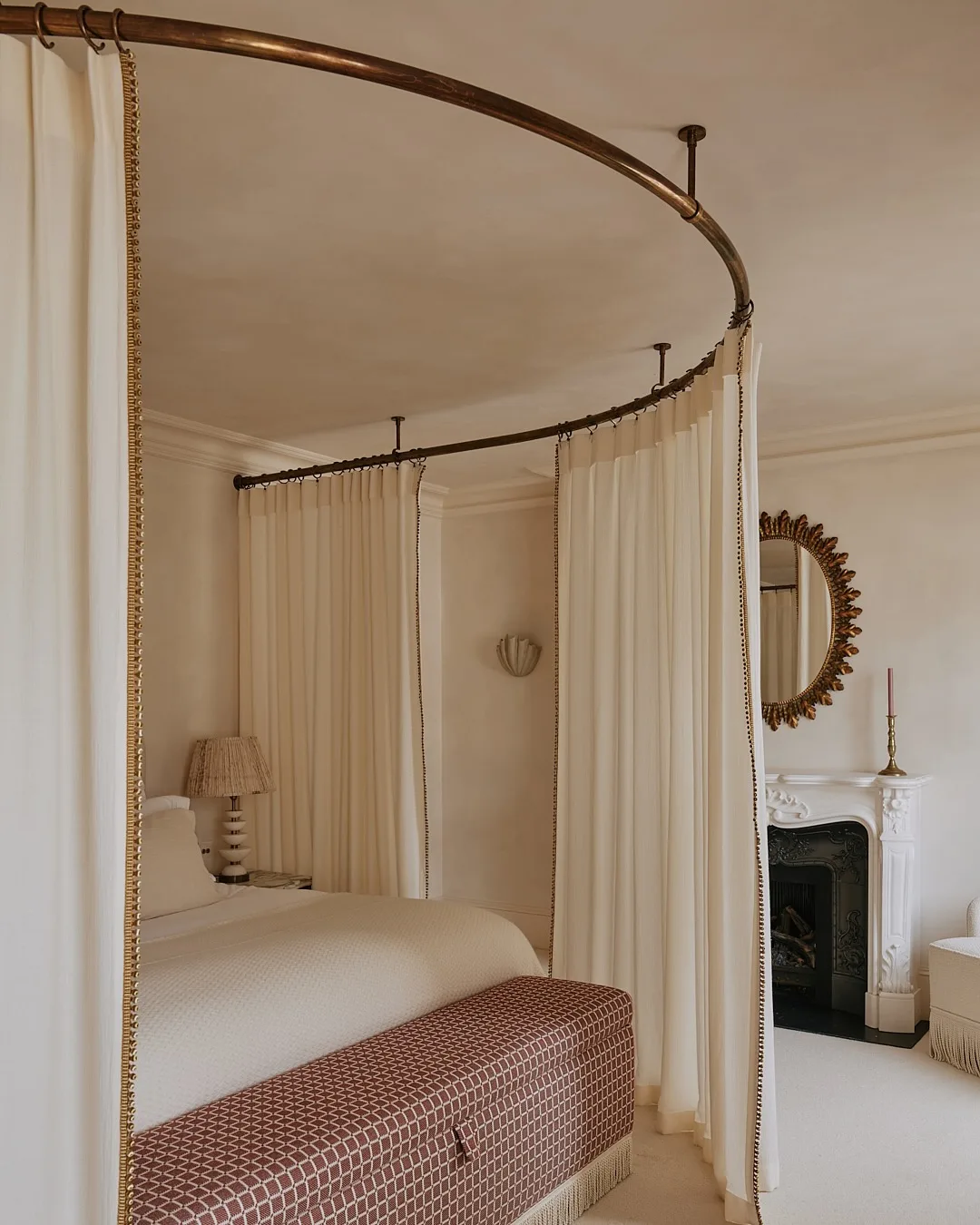 A cream bedroom with fireplace, mirror with gold frame, curtains around bed and red and white patterned foot stool.