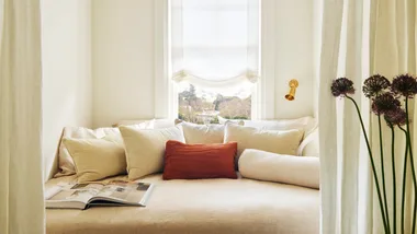 A bed sitting between two cream walls, white curtains natural linen bedding and red pillow