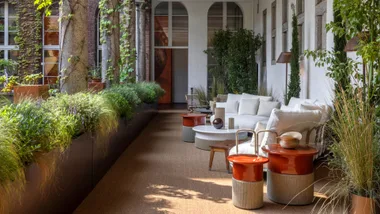 An alfresco balcony with white lounge furniture and orange and cord side tables flanked by greenery.