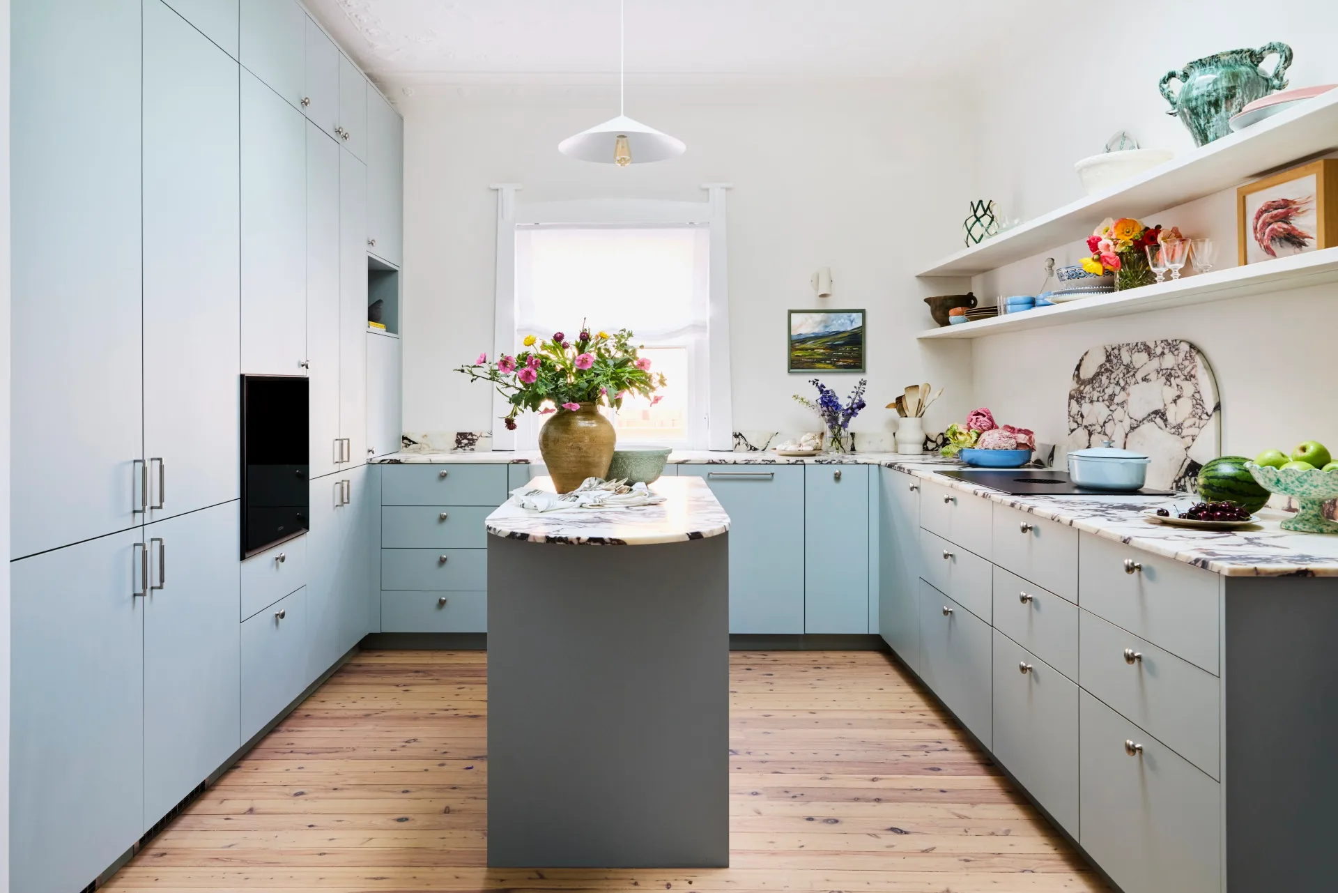 The blue home kitchen of Danielle Alvarez with narrow island bench surrounded by u-shaped blue cabinetry and white walls. 