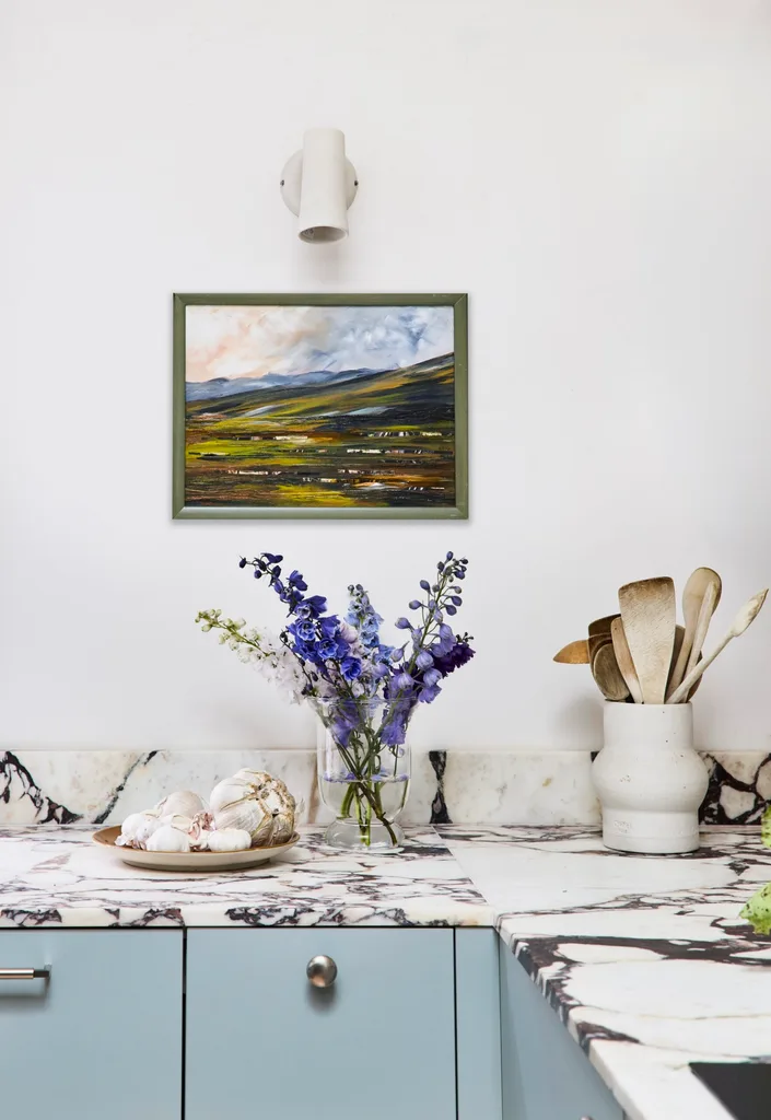A corner of Danielle Alvarez's home kitchen with a landscape painting on the wall below a white wall light with vessels and cooking utensils on the marble benchtop below.