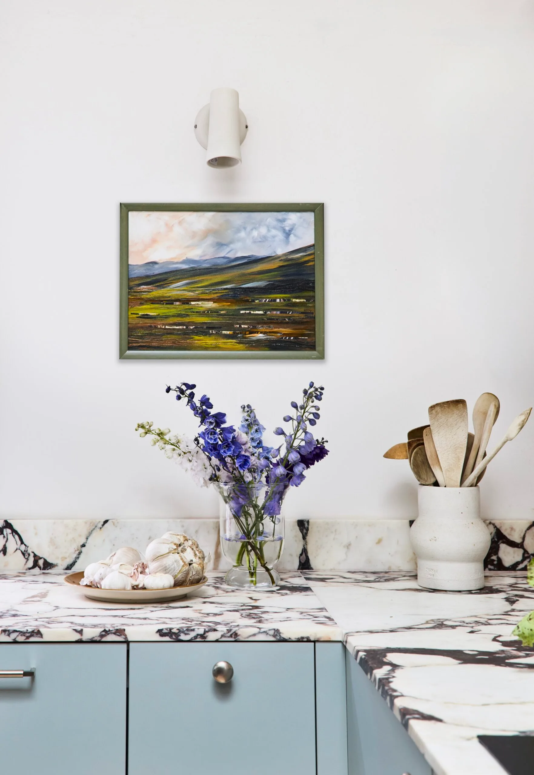 A corner of Danielle Alvarez's home kitchen with a landscape painting on the wall below a white wall light with vessels and cooking utensils on the marble benchtop below.