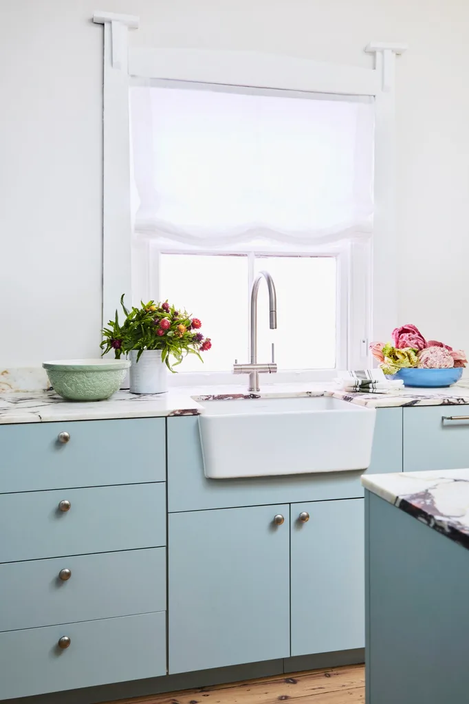 Danielle Alvarez home kitchen renovation blue cabinetry with a white farmhouse sink in front of a window with white roman blinds.