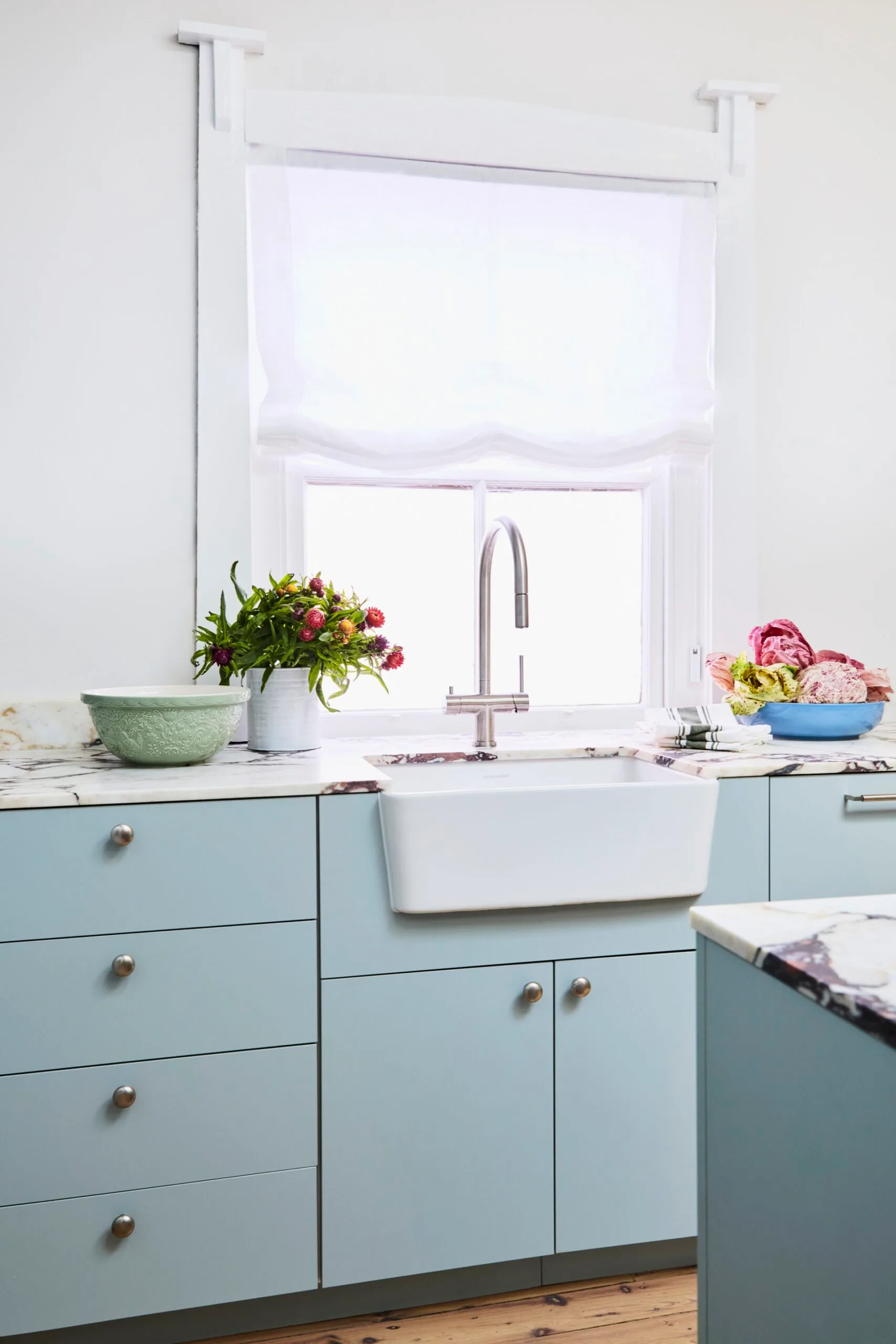 Danielle Alvarez home kitchen renovation blue cabinetry with a white farmhouse sink in front of a window with white roman blinds.
