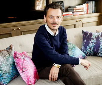 Man in a navy sweater and brown pants sits on a beige sofa with colorful pillows, wooden bookshelf in the background.