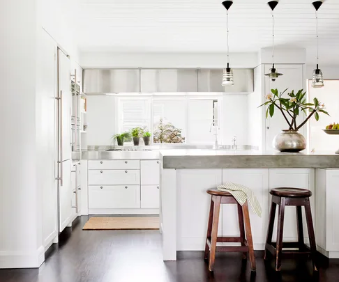 Modern white kitchen with stainless steel appliances, island, bar stools, hanging lights, and a vase with a plant on the counter.