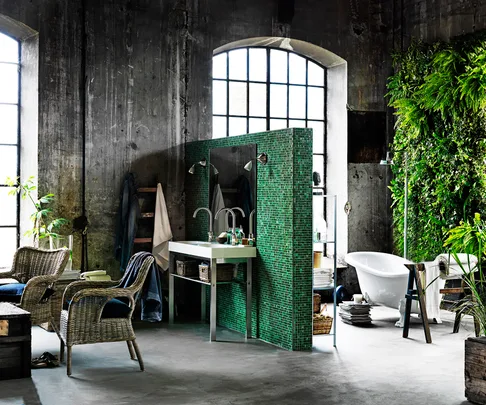 Industrial-style bathroom with green tiled partition, clawfoot tub, large windows, wicker chairs, and vertical plant wall.