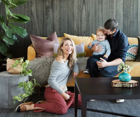 A smiling woman kneeling in front of a sofa, a man holding a cheerful toddler, with houseplants and colorful pillows around.
