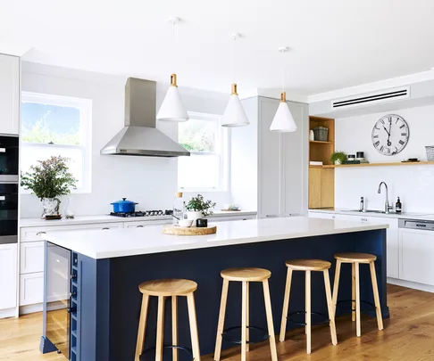 Modern kitchen with white countertops, navy island, wooden stools, pendant lights, and stainless steel appliances.