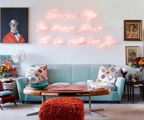 Mid-century styled living room with light blue sofa, eclectic pillows, neon wall art, vintage portraits, and various decorative items.
