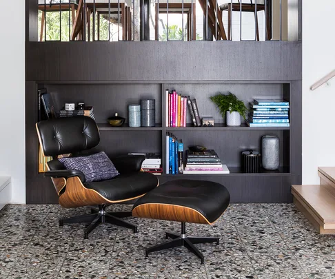 A black leather designer lounge chair with an ottoman in front of a dark wooden bookshelf filled with books and decorative items.