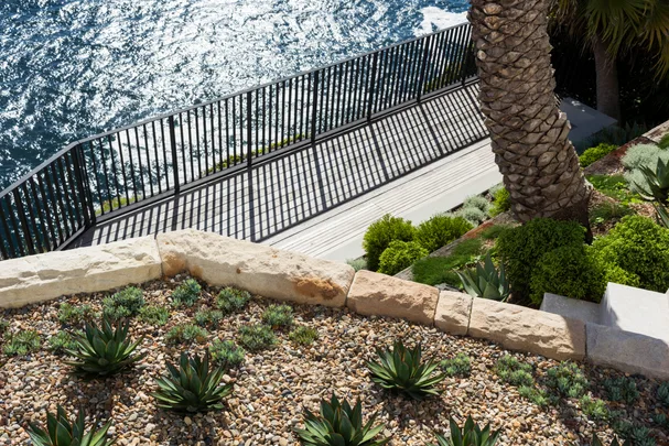 Clifftop garden with stone terrace, lush green plants, overlooking the ocean with metal railing and sparkling water below.