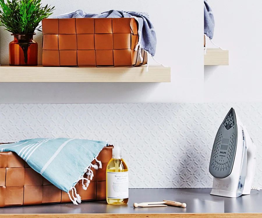 A laundry room shelf with a woven basket, green plant, blue towel, and an iron on a counter.
