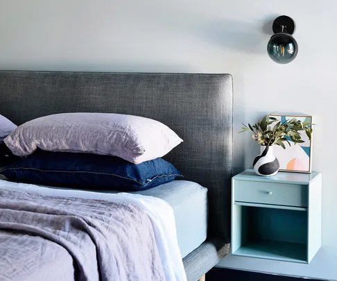 Bed with grey padded headboard, two stacked pillows, beside table with plant in vase, wall lamp above, minimal decor.