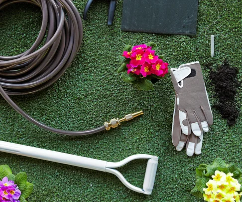 Gardening tools and gloves on grass with hose, flowers, and soil.