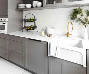 Modern kitchen with grey cabinets, a white countertop, gold faucet, farmhouse sink, and a basket of fruit.