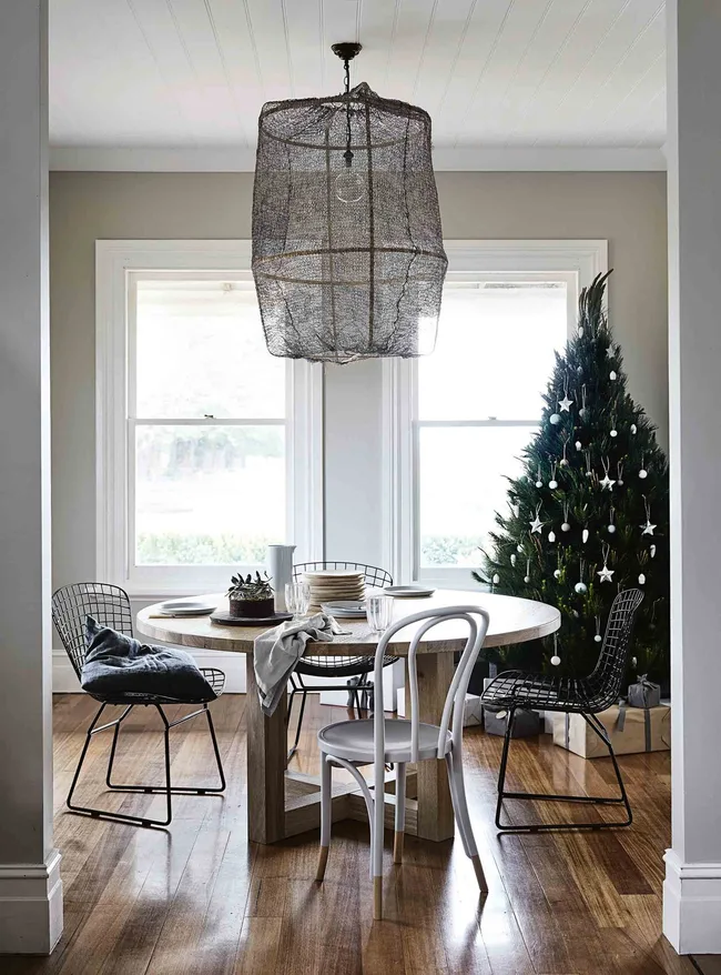 Dining room with a round table, four chairs, a wire chandelier, and a decorated Christmas tree by the window.