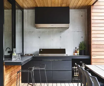 Modern outdoor kitchen with black cabinets, grill, beverages, and bar stools under wooden ceiling.