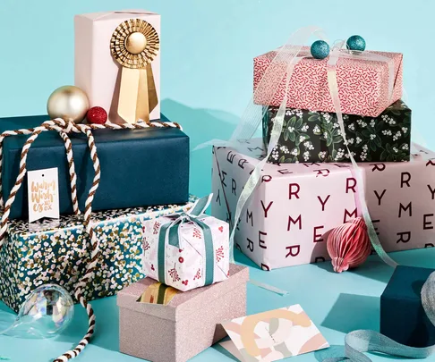 Colorfully wrapped Christmas presents stacked on a teal background, adorned with ribbons, ornaments, and minimalist gift tags.